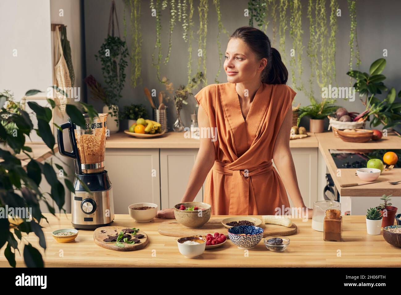 Happy young female in casual dress standing by kitchen table with various ingredients for homemade smoothie Stock Photo