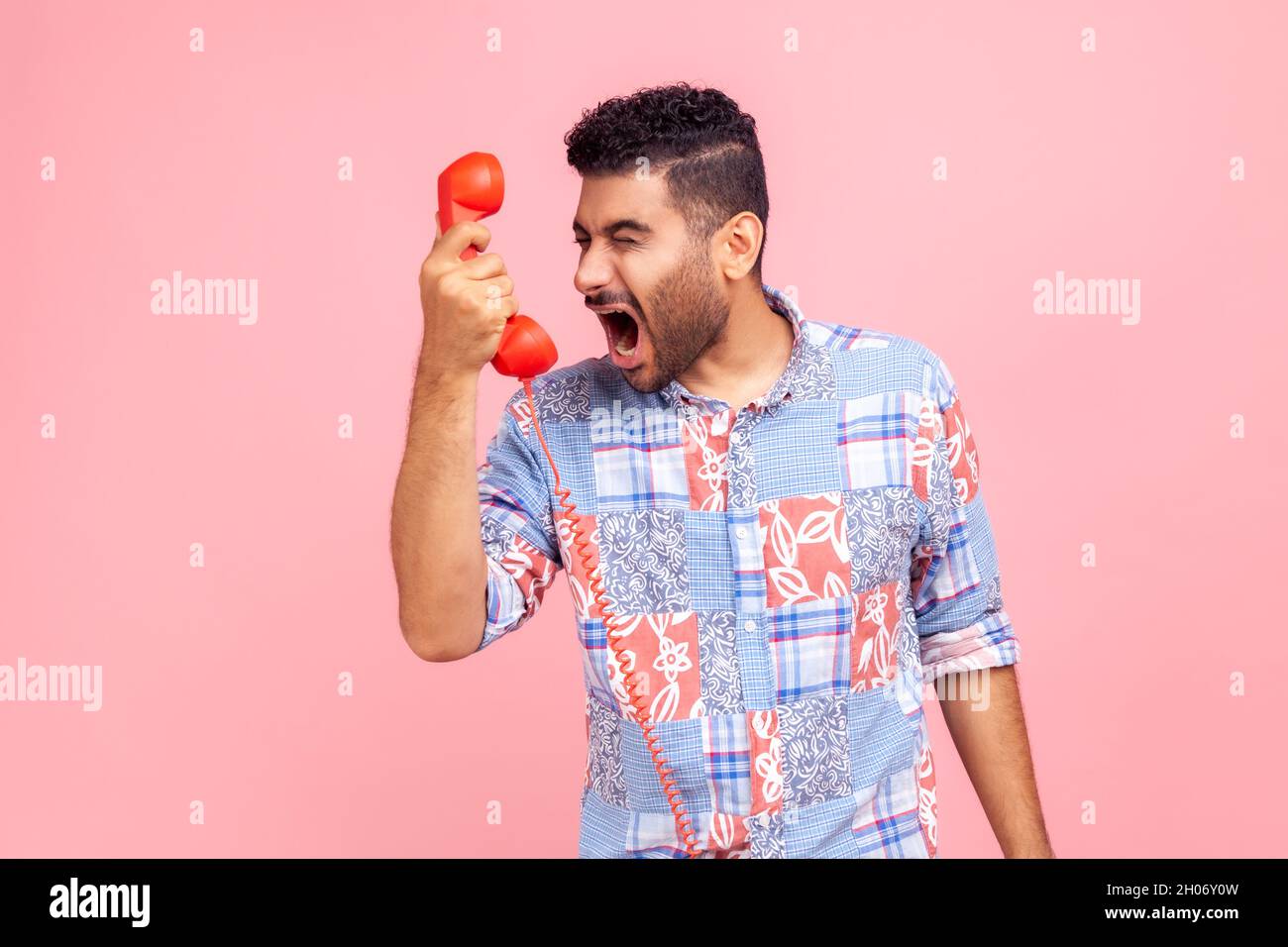 Portrait of bearded angry man screaming into phone handset, annoyed by telephone conversation, aggressive customer calling contact center. Indoor studio shot isolated on pink background. Stock Photo