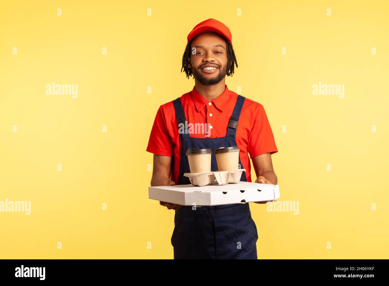 Delivery service. Friendly professional courier in blue overalls holding coffee and pizza box, wearing, offering drinks and food, looking at camera. Indoor studio shot isolated on yellow background. Stock Photo