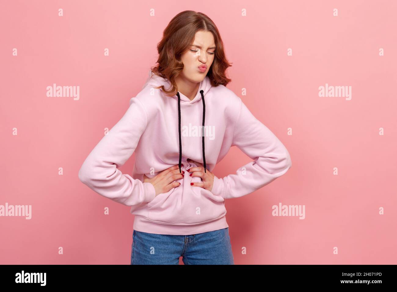 Portrait of upset teenager female in hoodie grimacing and holding belly, suffering abdominal pain and spasm, gastritis. Indoor studio shot isolated on pink background Stock Photo