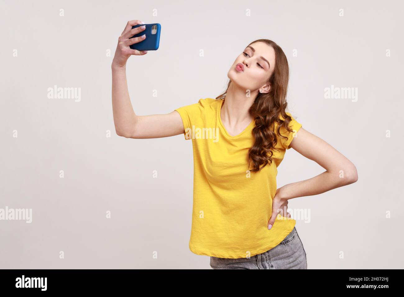 Portrait of cute curly haired teenage girl in yellow T-shirt sending kiss  on smartphone camera while communicating by video call or streaming vlog.  Indoor studio shot isolated on gray background Stock Photo -