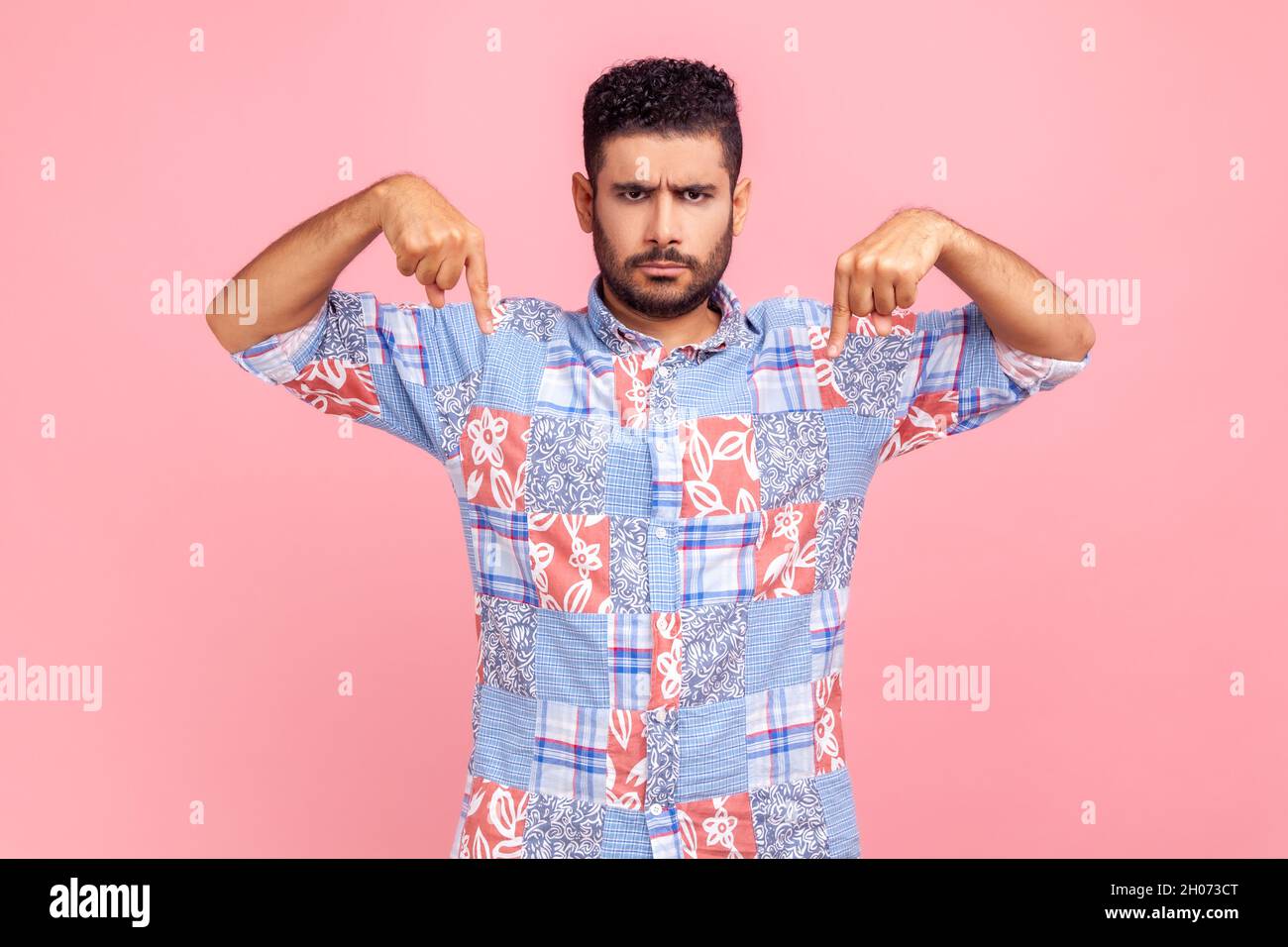 Here and now. Strict bearded handsome man pointing finger down, looking at camera with serious bossy facial expression, wearing blue casual shirt. Indoor studio shot isolated on pink background. Stock Photo