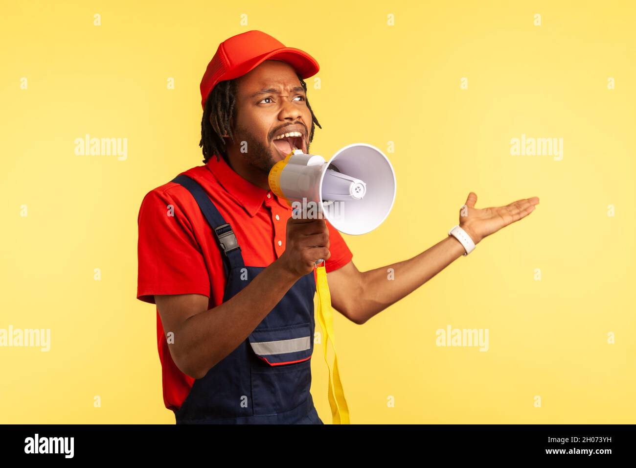 Attractive bearded handyman wearing red T-shirt and blue overalls holding megaphone, screaming, announcing discounts for service industry. Indoor studio shot isolated on yellow background. Stock Photo