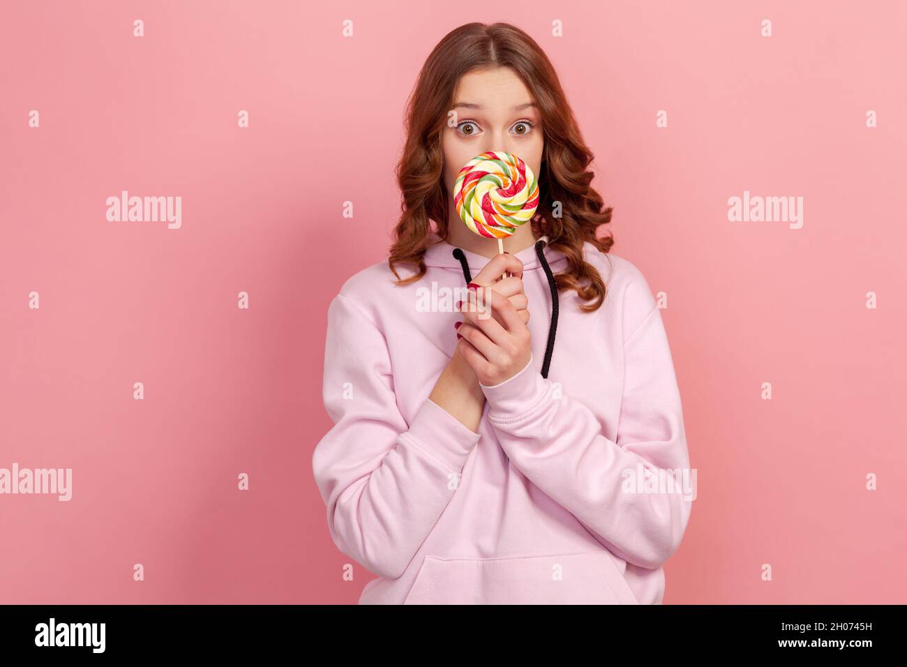 Portrait of surprised curly haired teenage girl in hoodie licking lollipop and looking at camera with amazed expression. Indoor studio shot isolated on pink background Stock Photo
