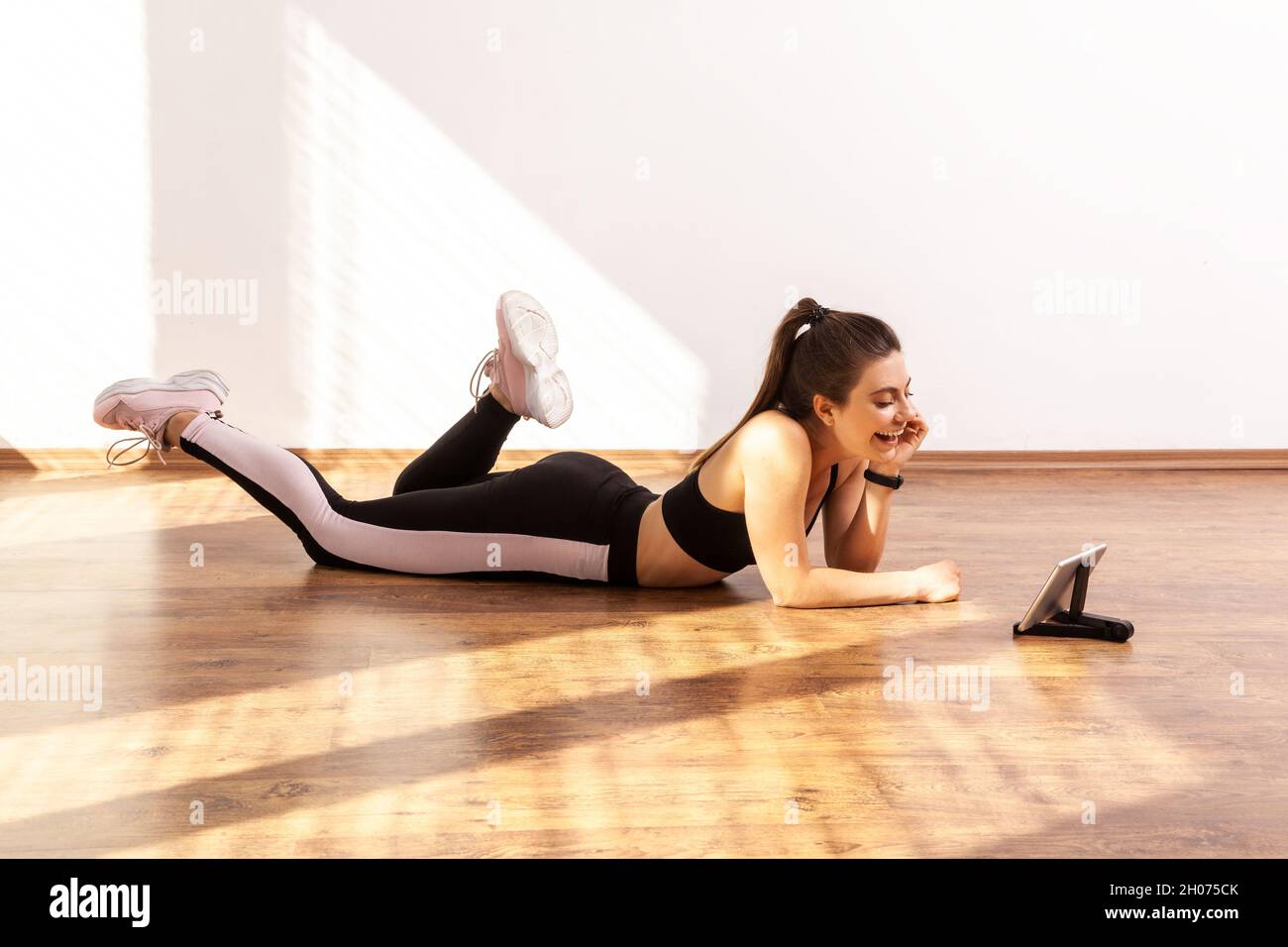 Slim female lying on floor and watching video about fitness exercising at home, looks happy, wearing black sports top and tights. Full length studio shot illuminated by sunlight from window. Stock Photo