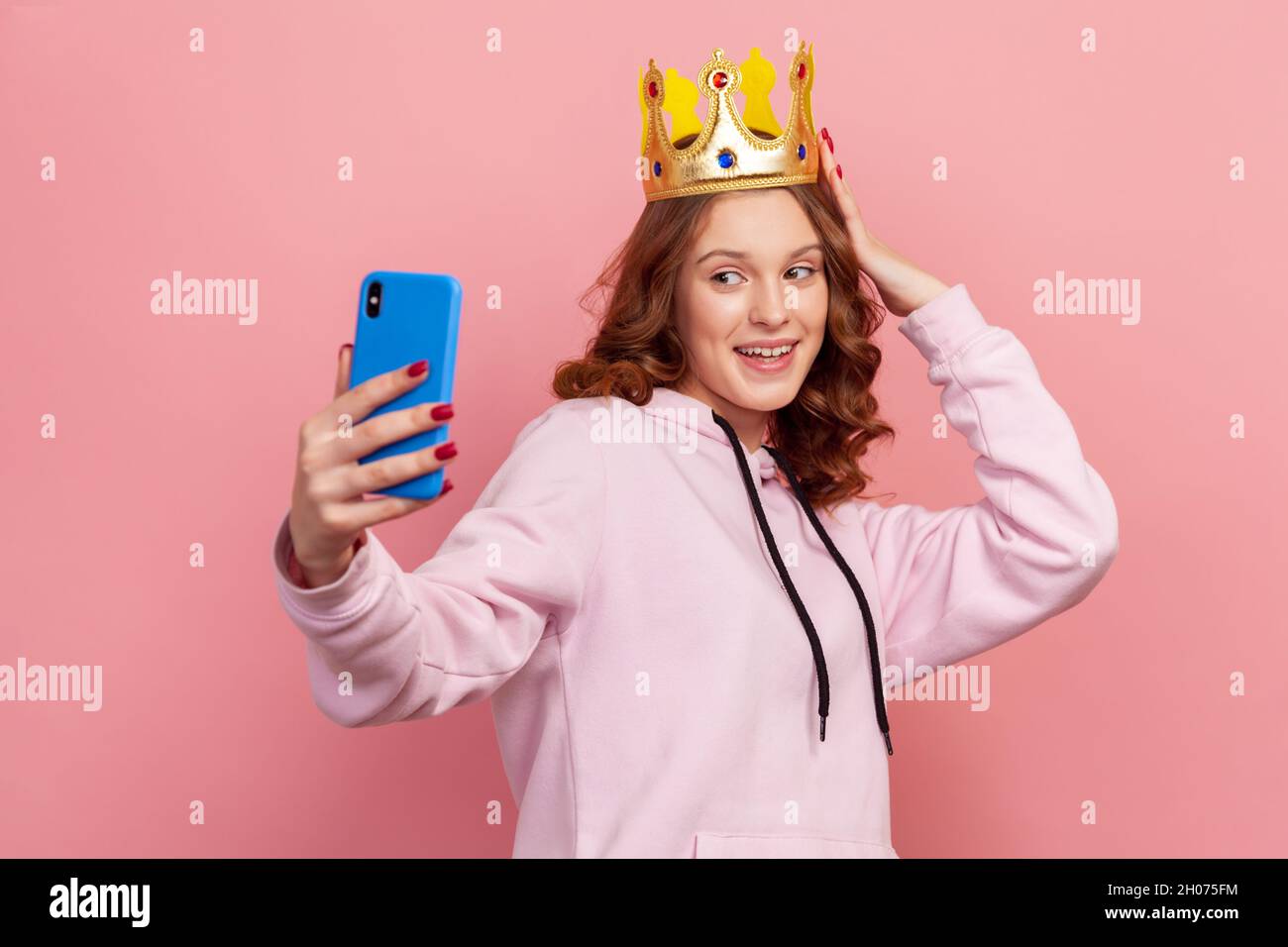 Portrait of smiling curly haired teenage girl in hoodie with gold crown posing on smartphone