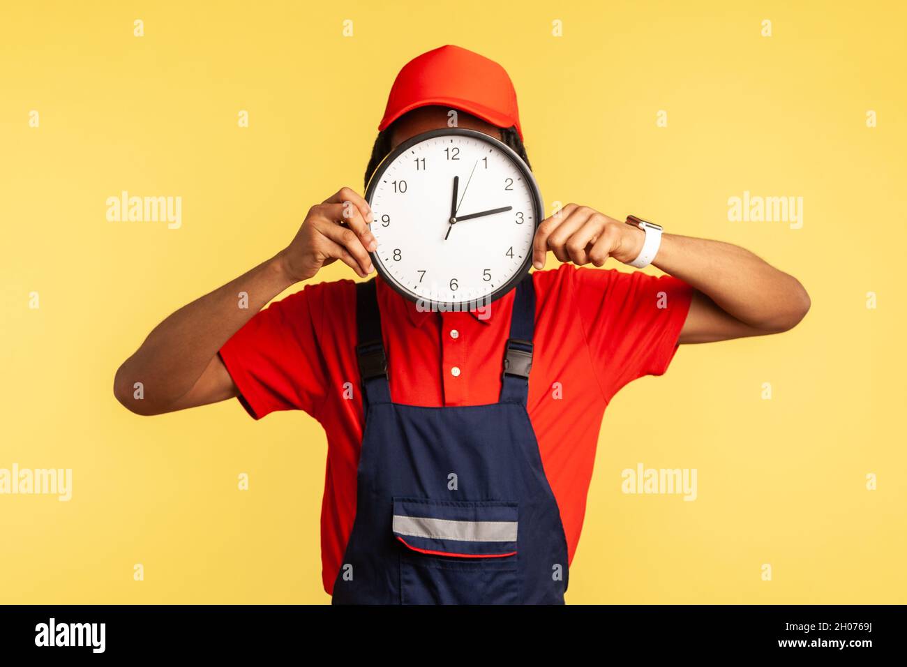 Anonymous labourer in uniform covering face with clock, advertising professional house repair services on time, punctual courier delivery. Indoor studio shot isolated on yellow background. Stock Photo