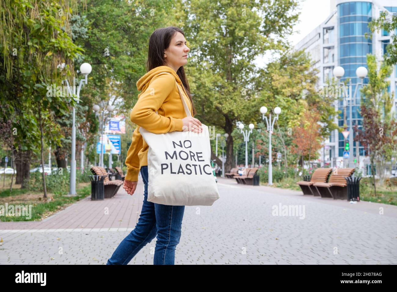 A brunette woman in an orange hoodie walks with a fabric bag that says No more plastic in the city Stock Photo