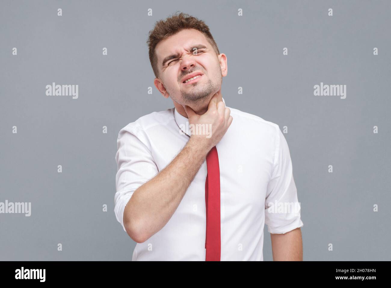 Throat pain. Portrait of young man in white shirt and tie standing and touching his painful neck. indoor isolated on gray background. Stock Photo