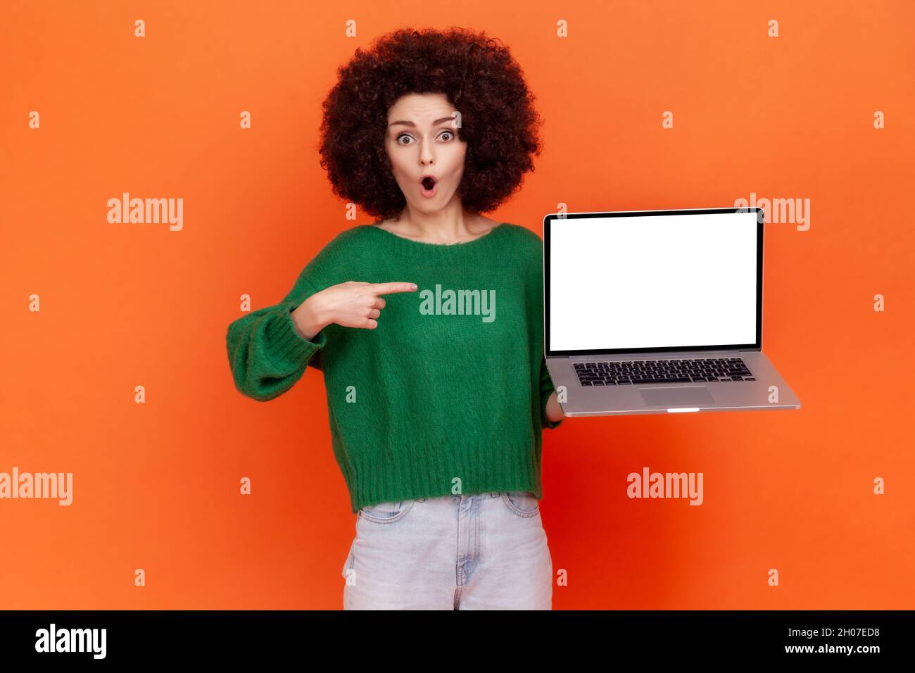Shocked woman with Afro hairstyle wearing green casual style sweater pointing at white empty display of her portable computer, copy space. Indoor studio shot isolated on orange background. Stock Photo
