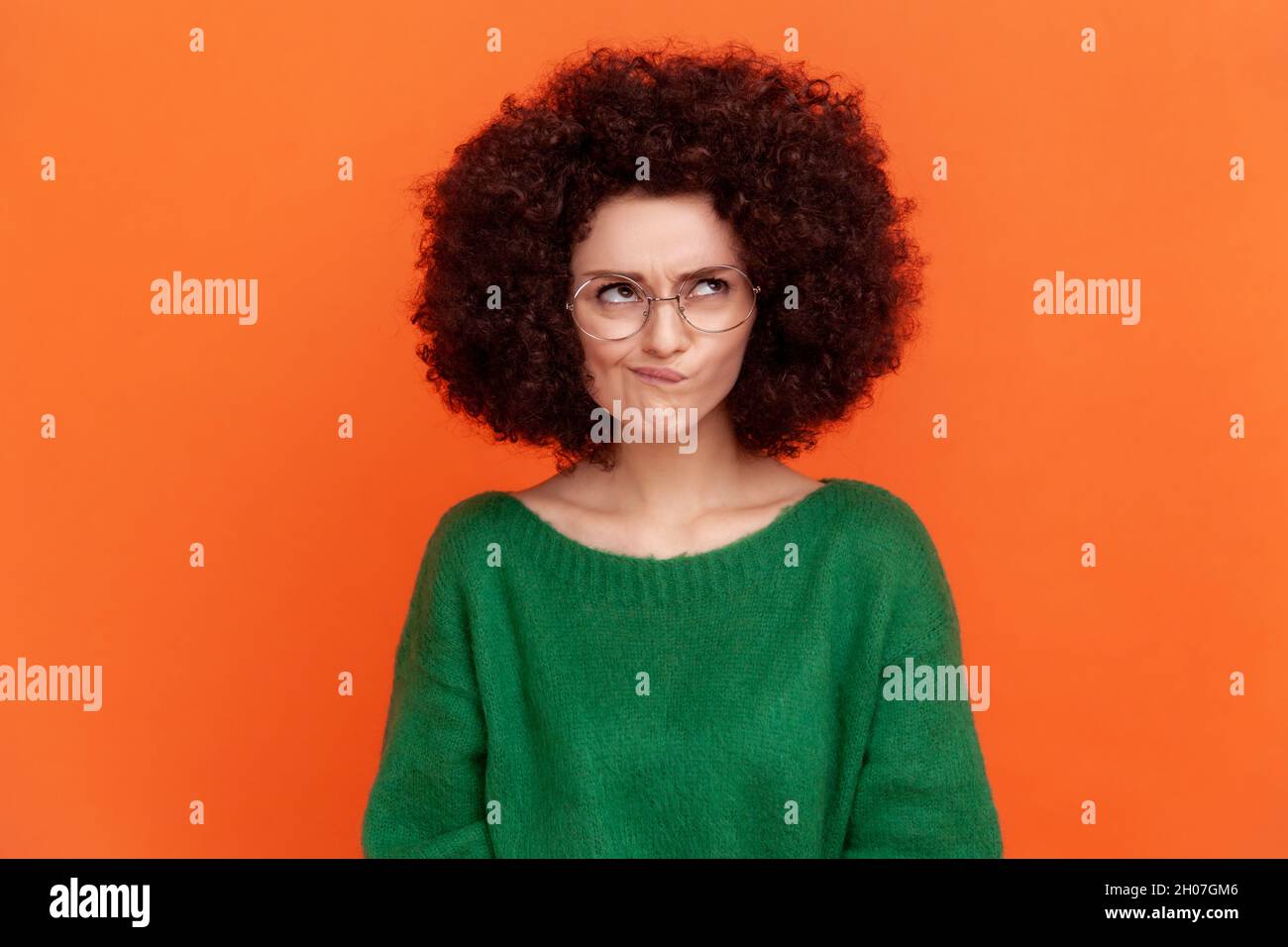 Portrait of confused woman with Afro hairstyle wearing green casual style sweater and optical glasses, looking away, frowning face, thinking. Indoor studio shot isolated on orange background. Stock Photo