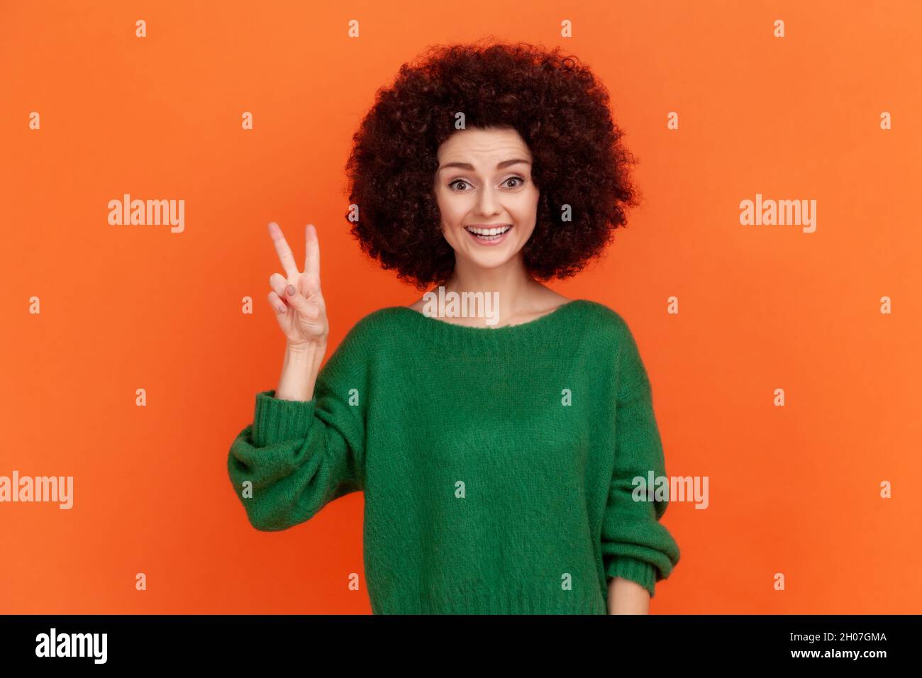 Happy friendly woman with Afro hairstyle wearing green casual style sweater looking at camera with toothy smile, showing v sign, peace gesture. Indoor studio shot isolated on orange background. Stock Photo