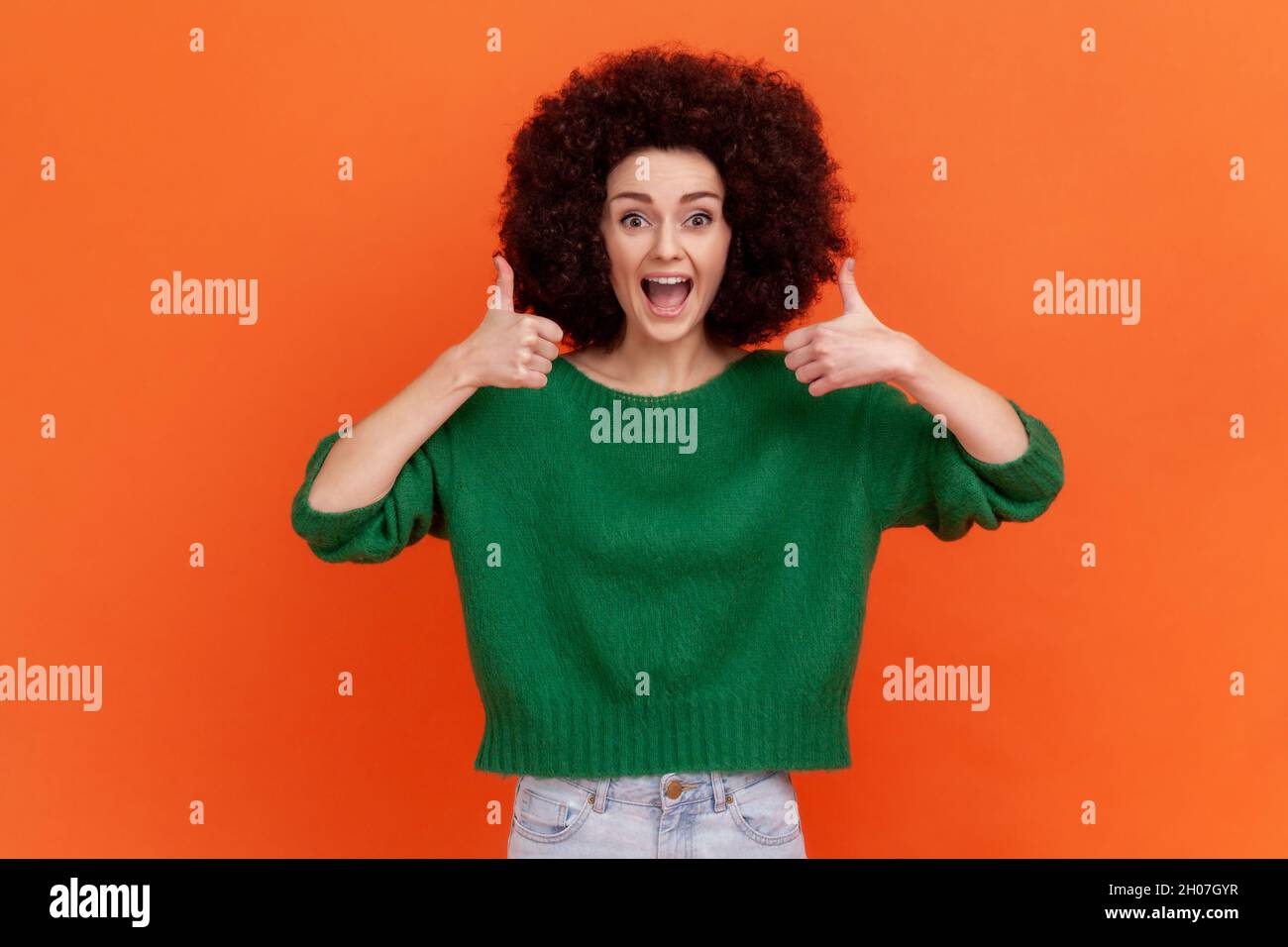 Amazed positive woman with Afro hairstyle wearing green casual style sweater standing showing thumb up with both hands, recommend content. Indoor studio shot isolated on orange background. Stock Photo