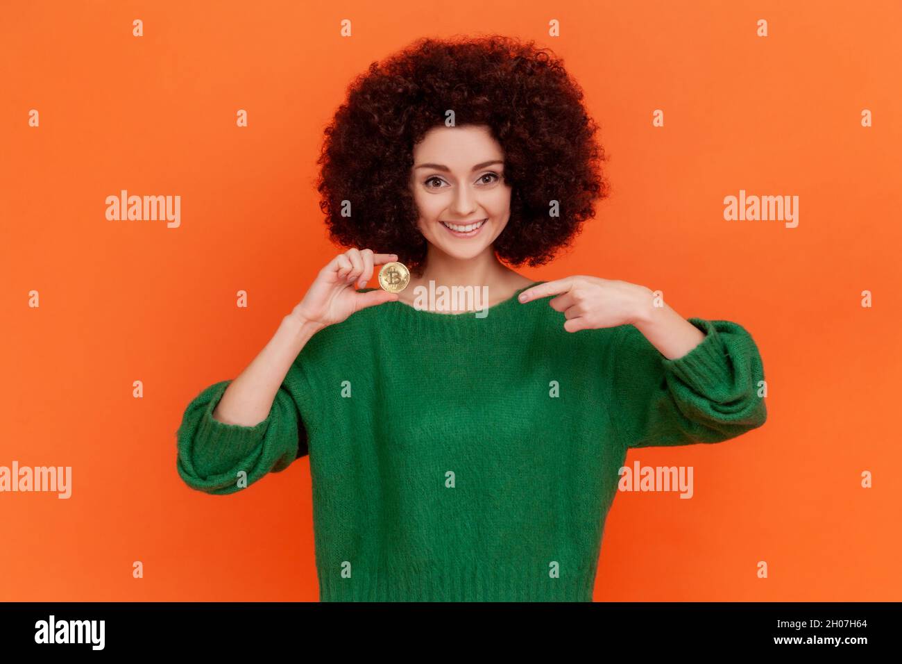 Portrait of pleasant looking woman with Afro hairstyle wearing green casual style sweater pointing finger at gold bitcoin, ecommerce. Indoor studio shot isolated on orange background. Stock Photo