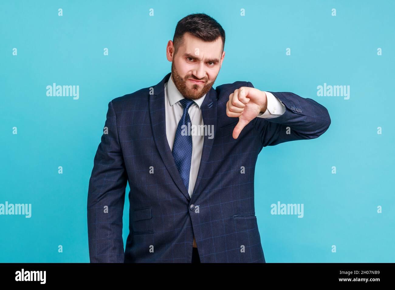 Portrait of unsatisfied bearded man wearing official style suit showing thumb down and looking at camera, dislike, application testing feedback. Indoor studio shot isolated on blue background. Stock Photo