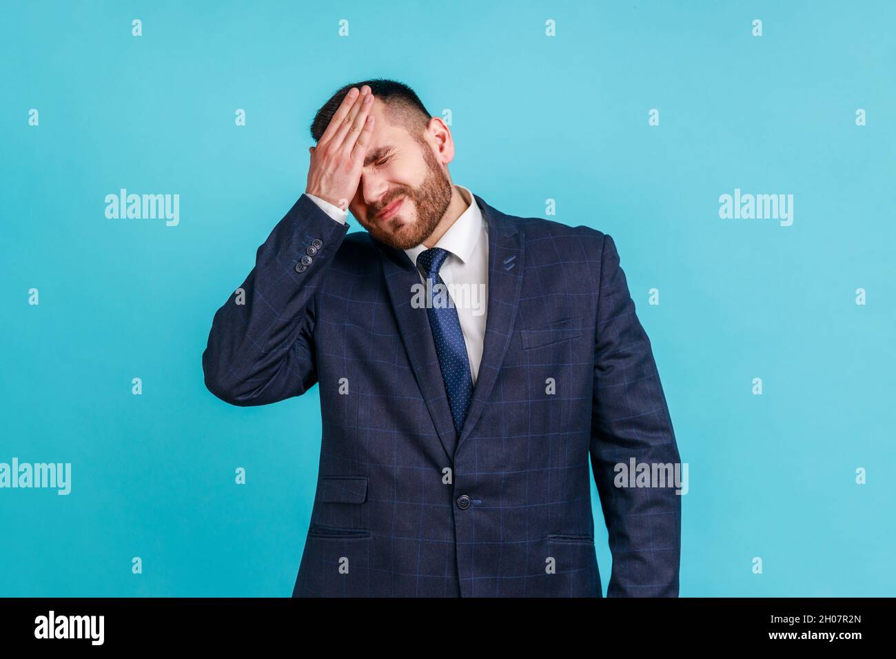 Portrait of upset bearded man wearing official style suit standing with facepalm gesture, blaming himself, feeling sorrow regret because of bad memory. Indoor studio shot isolated on blue background. Stock Photo