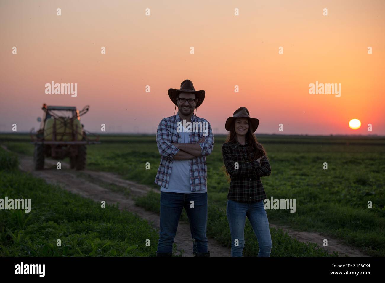 Two farmers standing with crossed arms in front of tractor at sunset ...