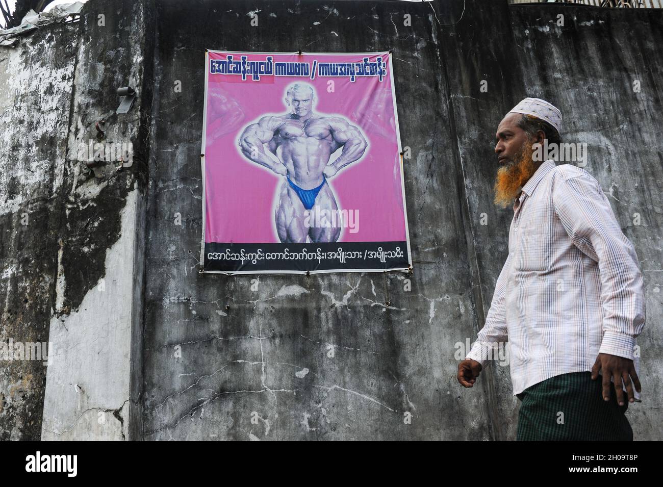 '11.10.2013, Myanmar, , Yangon - A Muslim man wearing a traditional takke walks past a wall with a poster advertising a bodybuilding event. 0SL131011D Stock Photo