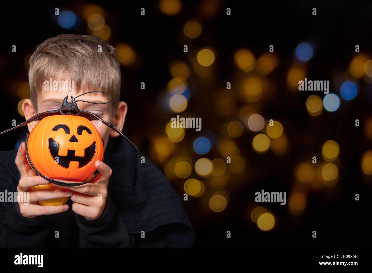 A child holding an orange pumpkin-shaped basket with a grinning face, Jack's lantern and a bat on a black background with bokeh. The boy is waiting fo Stock Photo