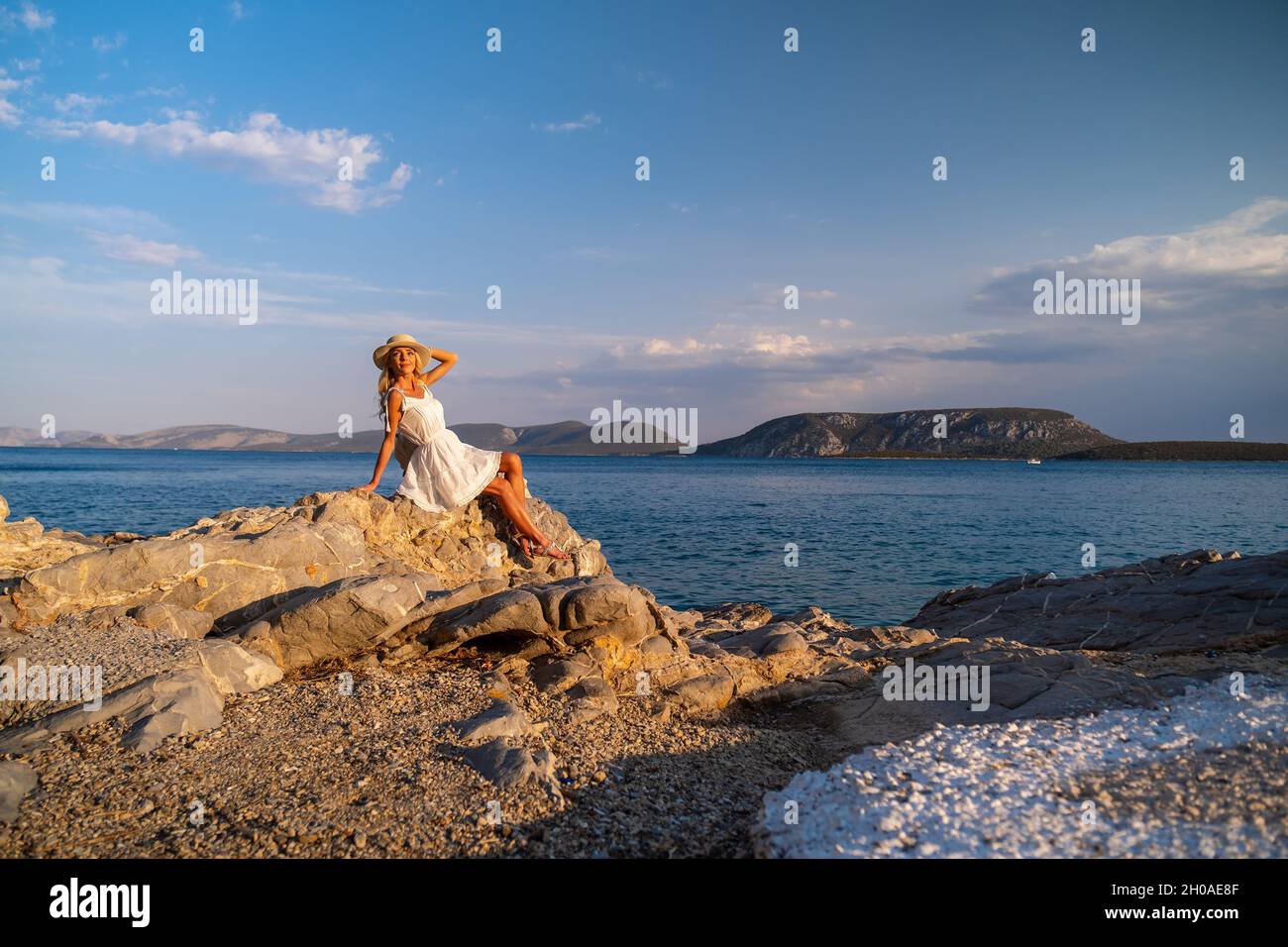 Beautiful young woman wearing traw hat sitting in rocks in white dress by the seashore Stock Photo