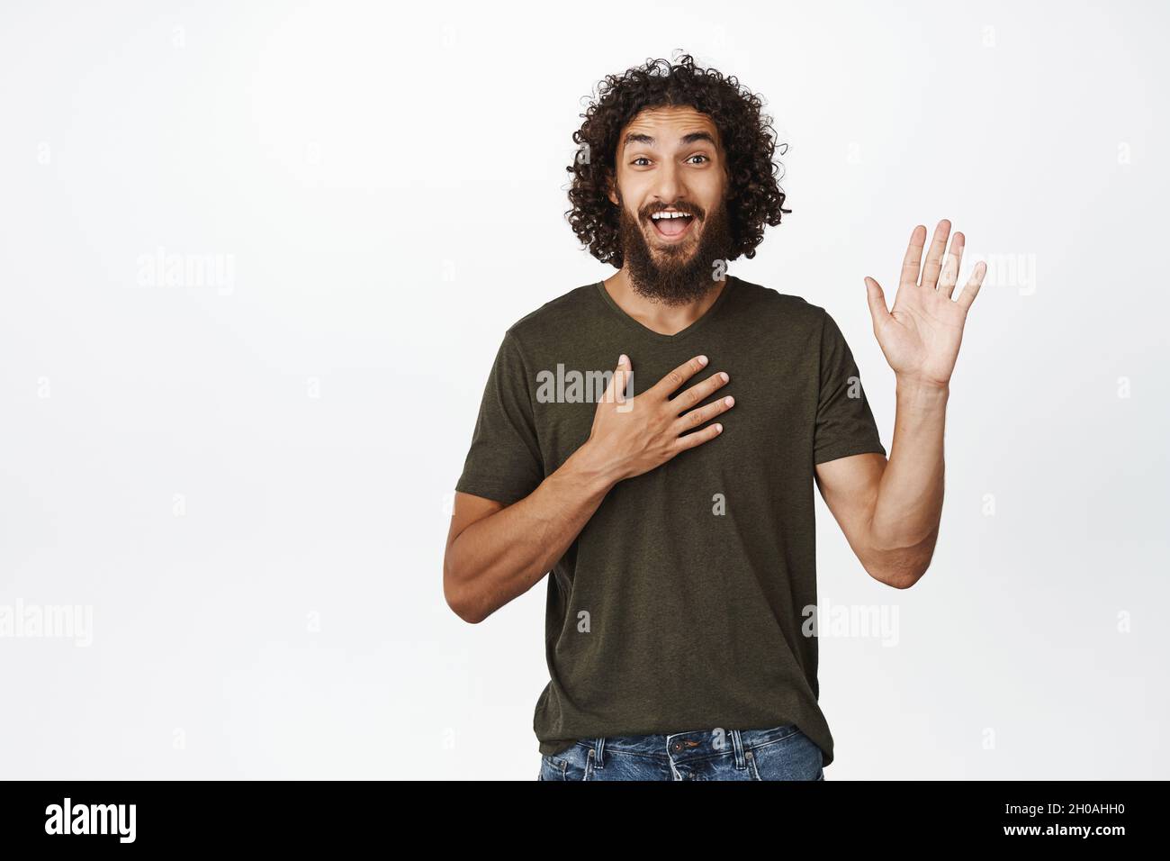 Enthusiastic bearded guy smiling, raising one arm and put hand on heart, introduce himself, waving to greet you, standing over white background Stock Photo