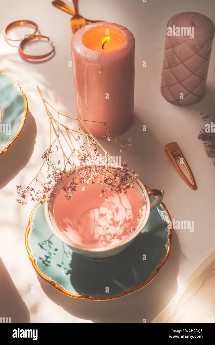 Close up of pink flowers tea in blue porcelain tea cup on white table with burning candle with golden jewelry. Cozy lifestyle concept. Top view. Stock Photo