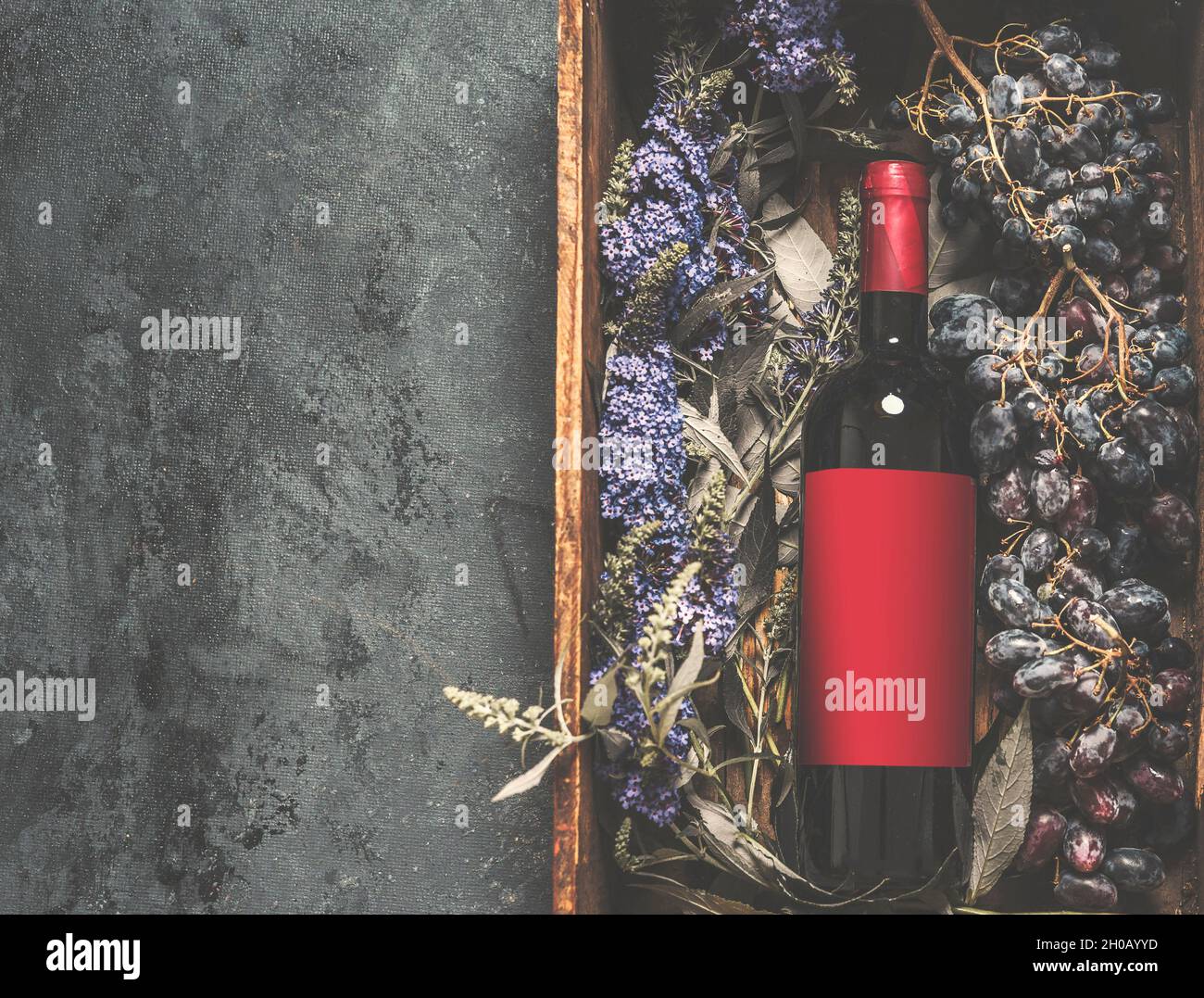 Red wine bottle with empty red label mock up with copy space in wooden box with grapes and flowers on dark concrete kitchen table. Rustic red wine con Stock Photo