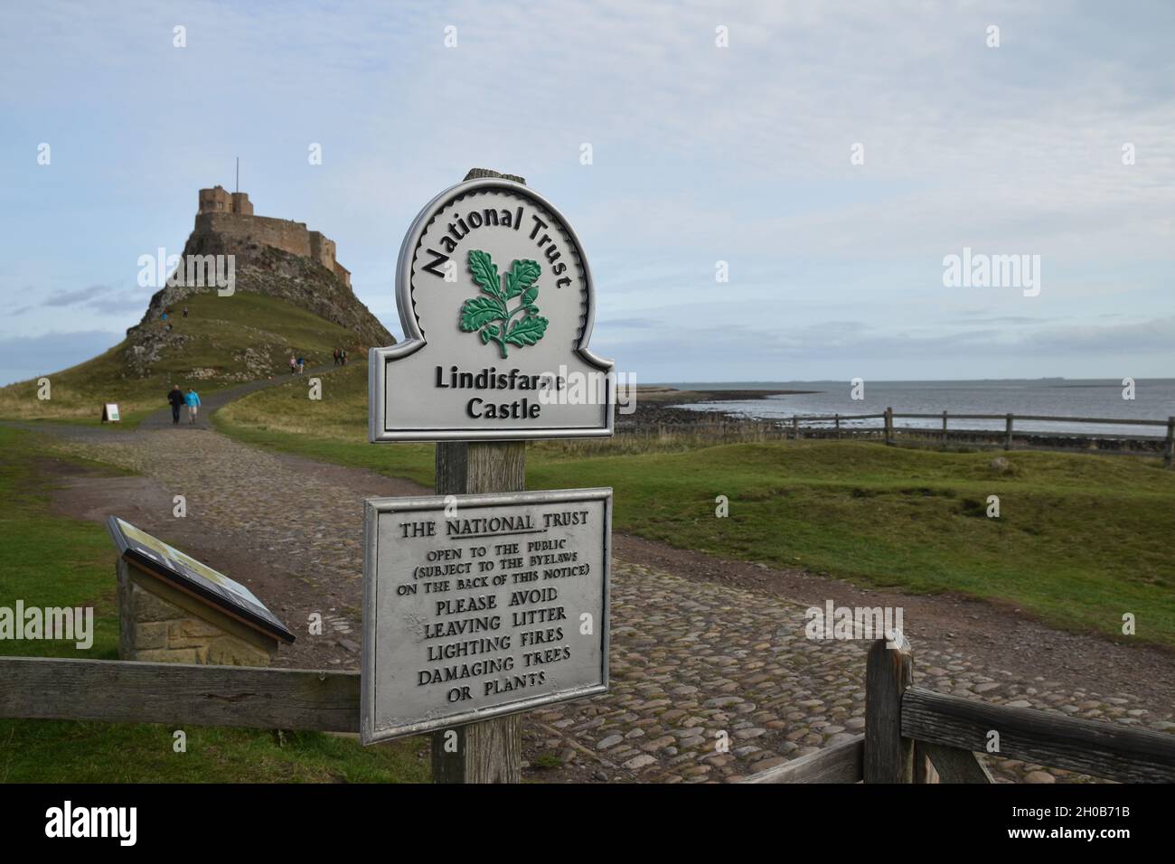 Sign for Lindisfarne Castle with blurred background of path, castle and blue sky with clouds. Shallow depth of field. Stock Photo