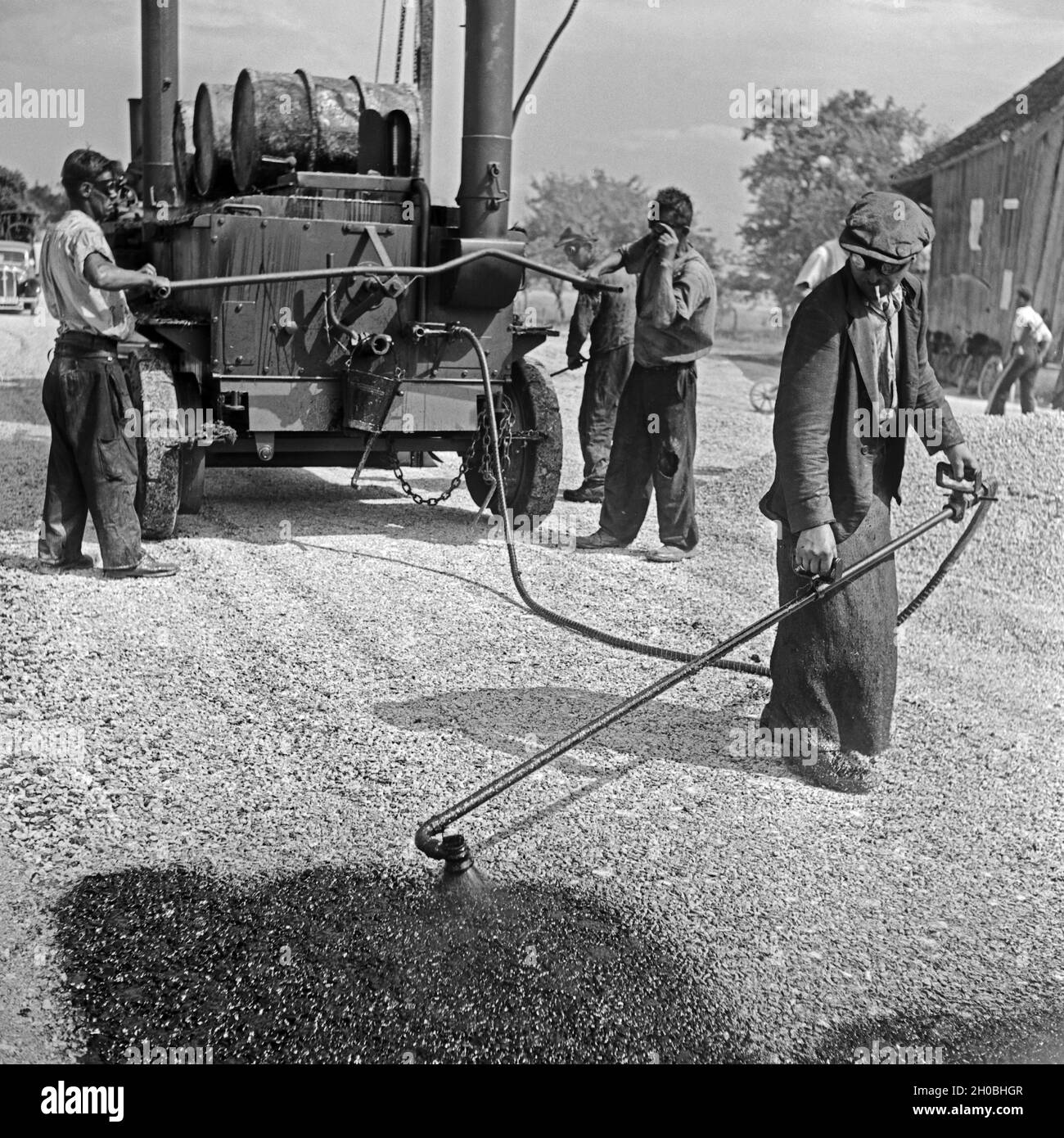 Straßenbauarbeiter an einer Teerküche bei Straßenbauarbeiten in der Nähe von Burghausen, einer erhitzt den Straßenbelag, Deutschland 1930er Jahre. Construction workers working at a construction area with a ropemaker's stove, one of them scalding the tarmac near Burghausen, Germany 1930s. Stock Photo