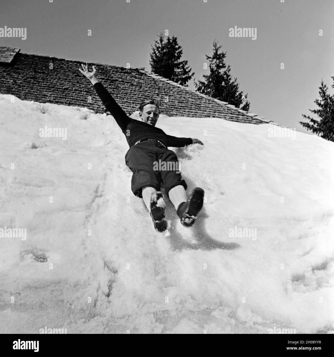 Ein fröhlicher Skiurlauber rutscht auf dem Hosenboden das verschneite Hoteldach hinunter, Deutschland 1930er Jahre. A happy ski tourist gliding down from the snowed in roof of the hotel, Germany 1930s. Stock Photo