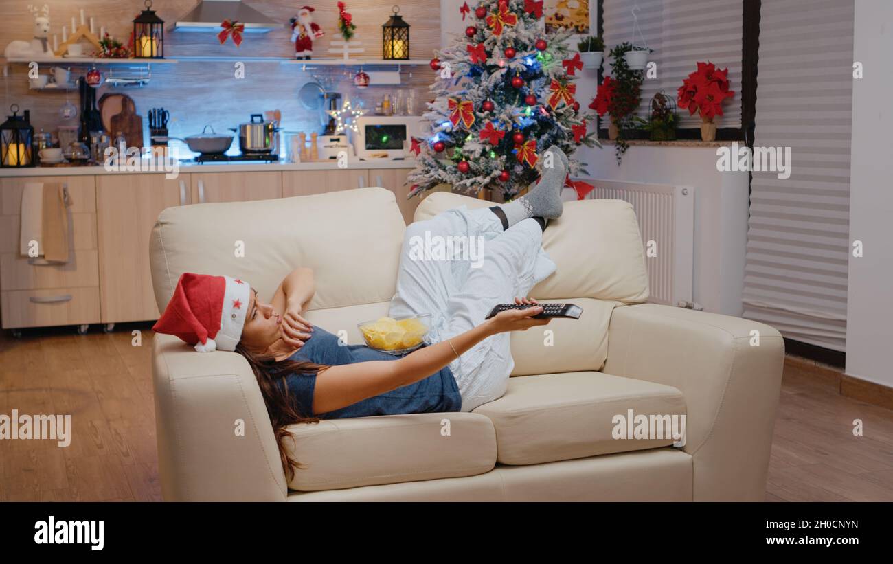 Bored adult switching channels with TV remote control while laying on  couch. Woman wearing santa hat and watching television, eating chips from  bowl on christmas eve festivity Stock Photo - Alamy