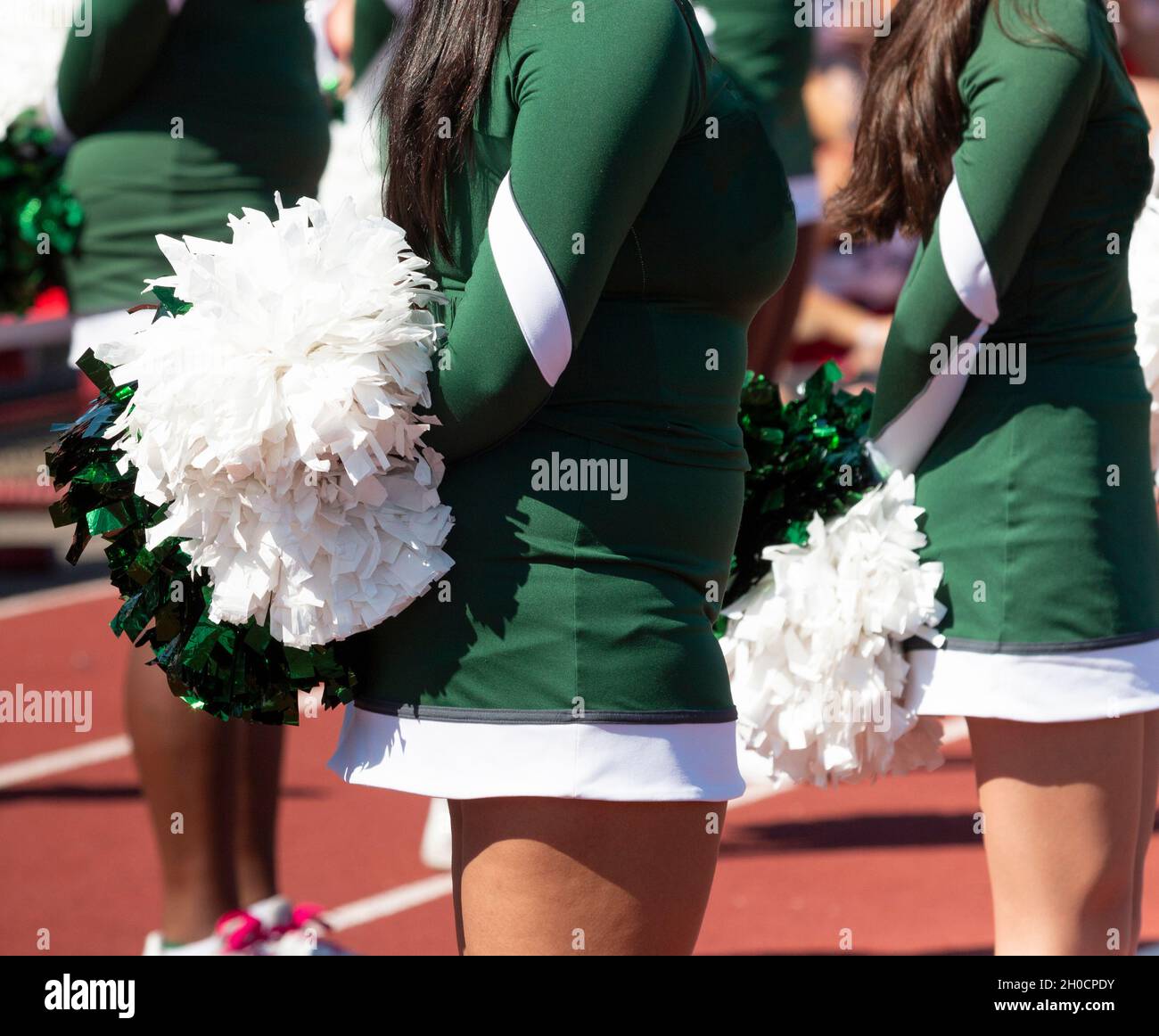 Close up of high school cheerleaders in green uniforms holding white and green pom poms behind their backs. Stock Photo