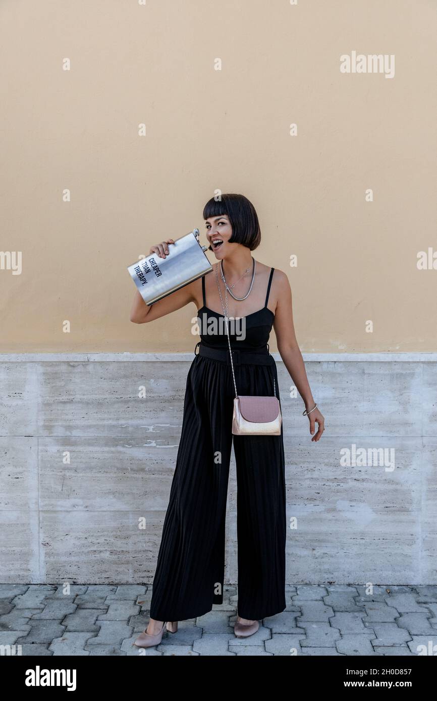 Portrait of happy young woman wearing elegant black jumpsuit, holding large liquor flask Stock Photo