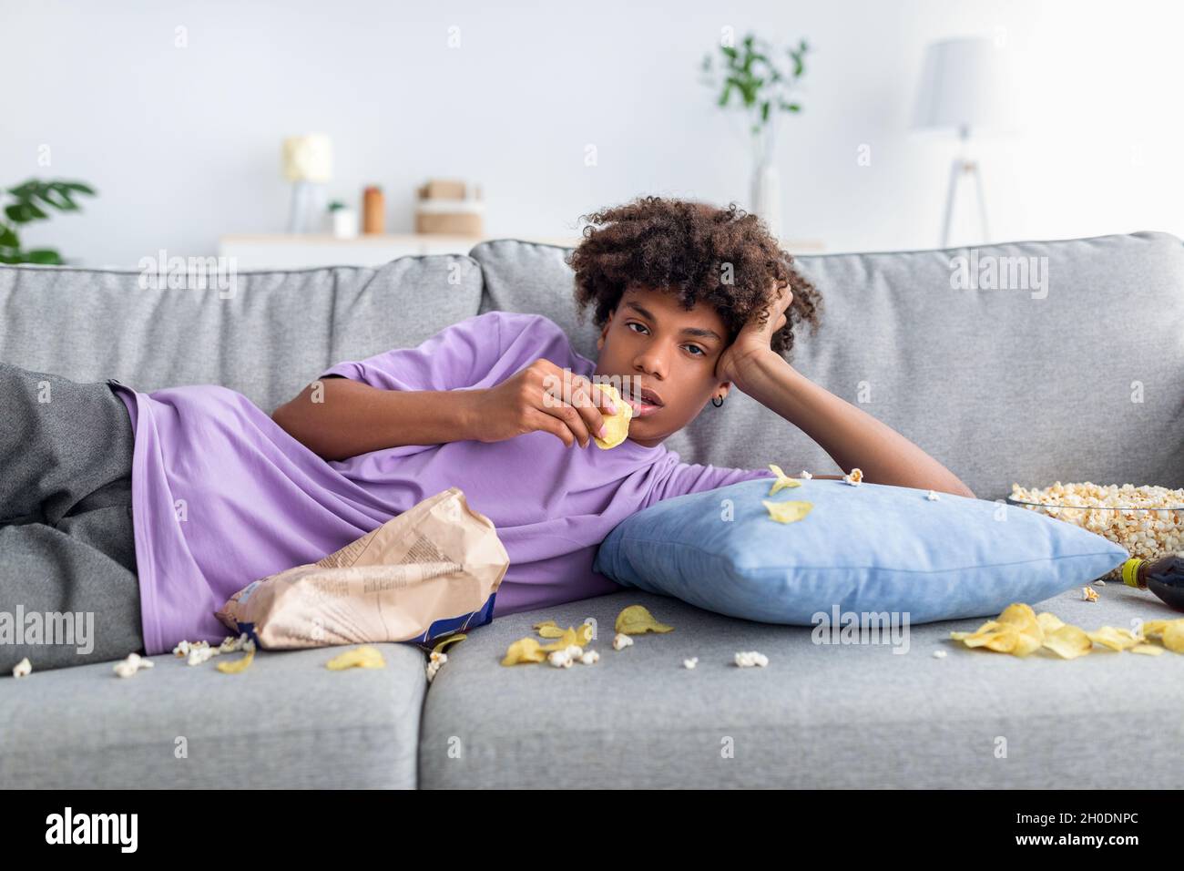 Bored Afro teenager lying on sofa with scattered snacks, eating chips,  watching dull show or movie on TV at home Stock Photo - Alamy