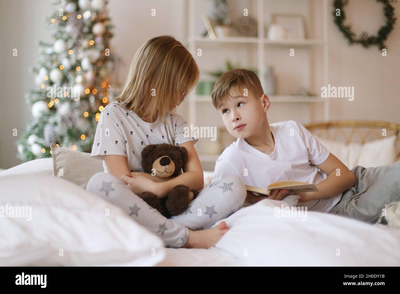 Brother is reading a book to his sister, sitting on the bed. Stock Photo