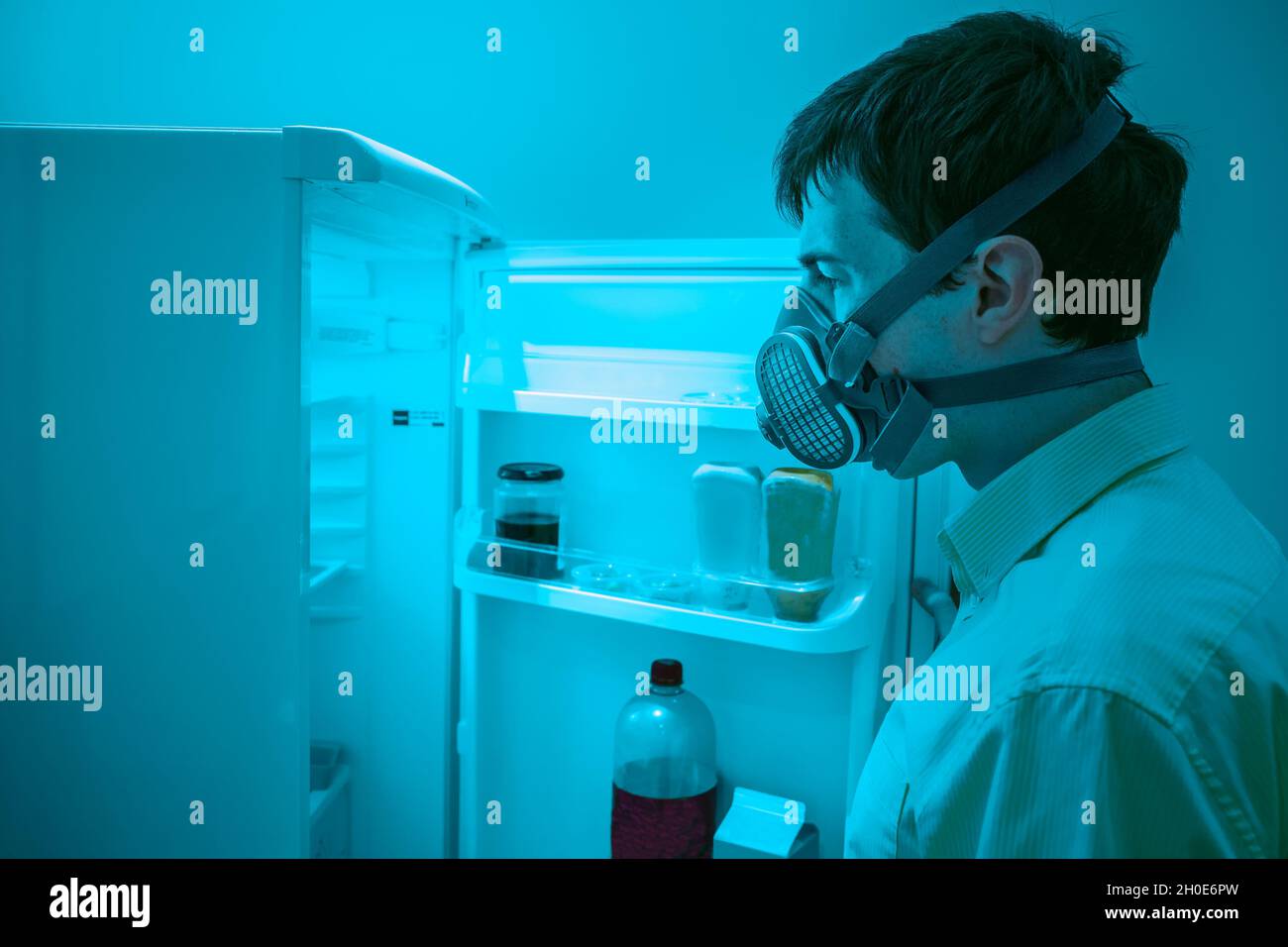 A man wearing an air filtration mask opens a refridgerator door. Perhaps he is concerned about what he may find inside. Stock Photo