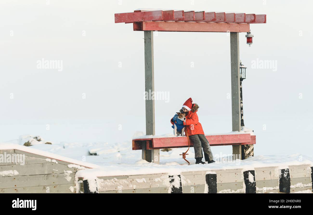 Kid boy wearing Santa Claus hat warming up his pet dog sitting on bench on pier with frozen white sea in background Stock Photo