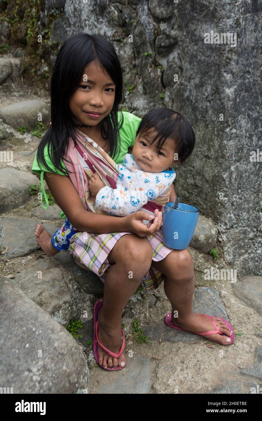 Young Filipino girl holding her little sister on her lap while feeding her. Batad village, Ifugao province, Philippines Stock Photo