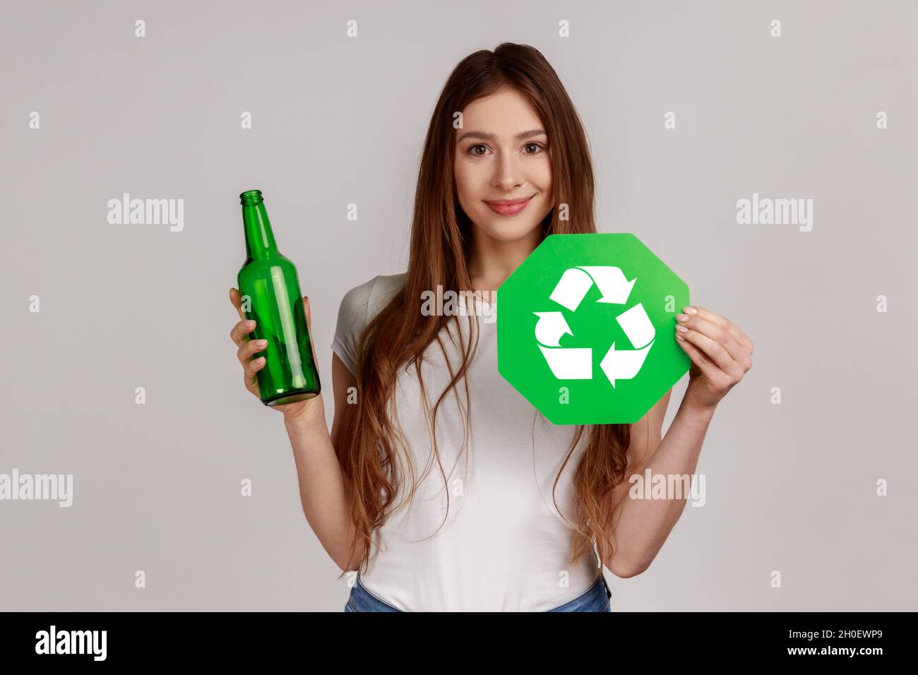 Woman holding glass bottle and green recycling sign, garbage sorting and environment protection, wearing white casual style T-shirt. Indoor studio shot isolated on gray background. Stock Photo