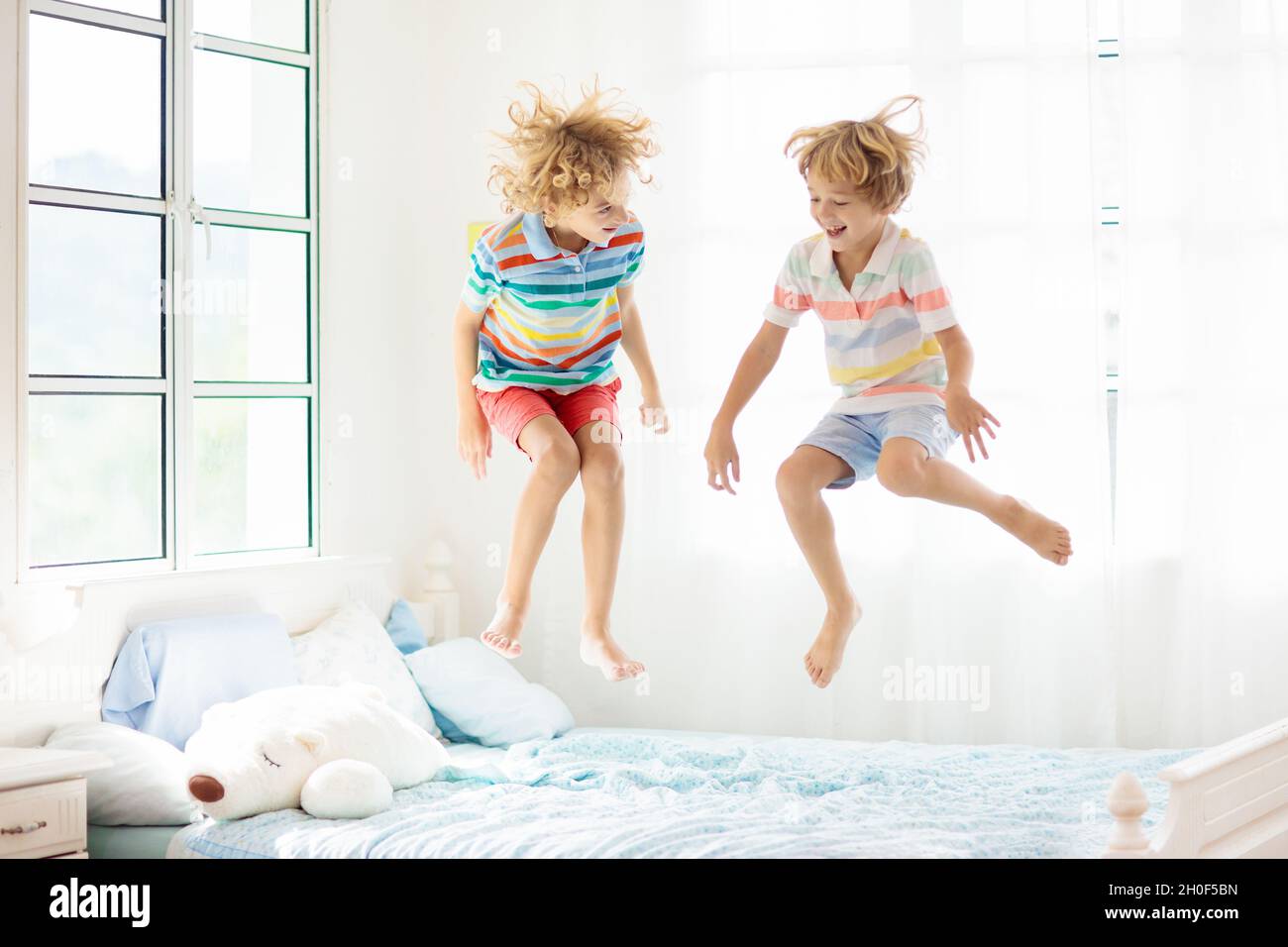 Child jumping on bed in white bedroom with big window. Kids play at home. Fun jump on parents