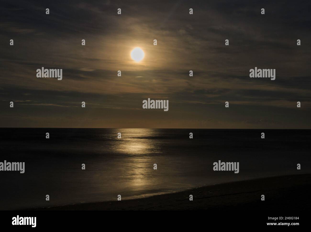 Long exposure, night beach scene with large full moon and clouds sitting over a calm, smooth sea and surf. Stock Photo