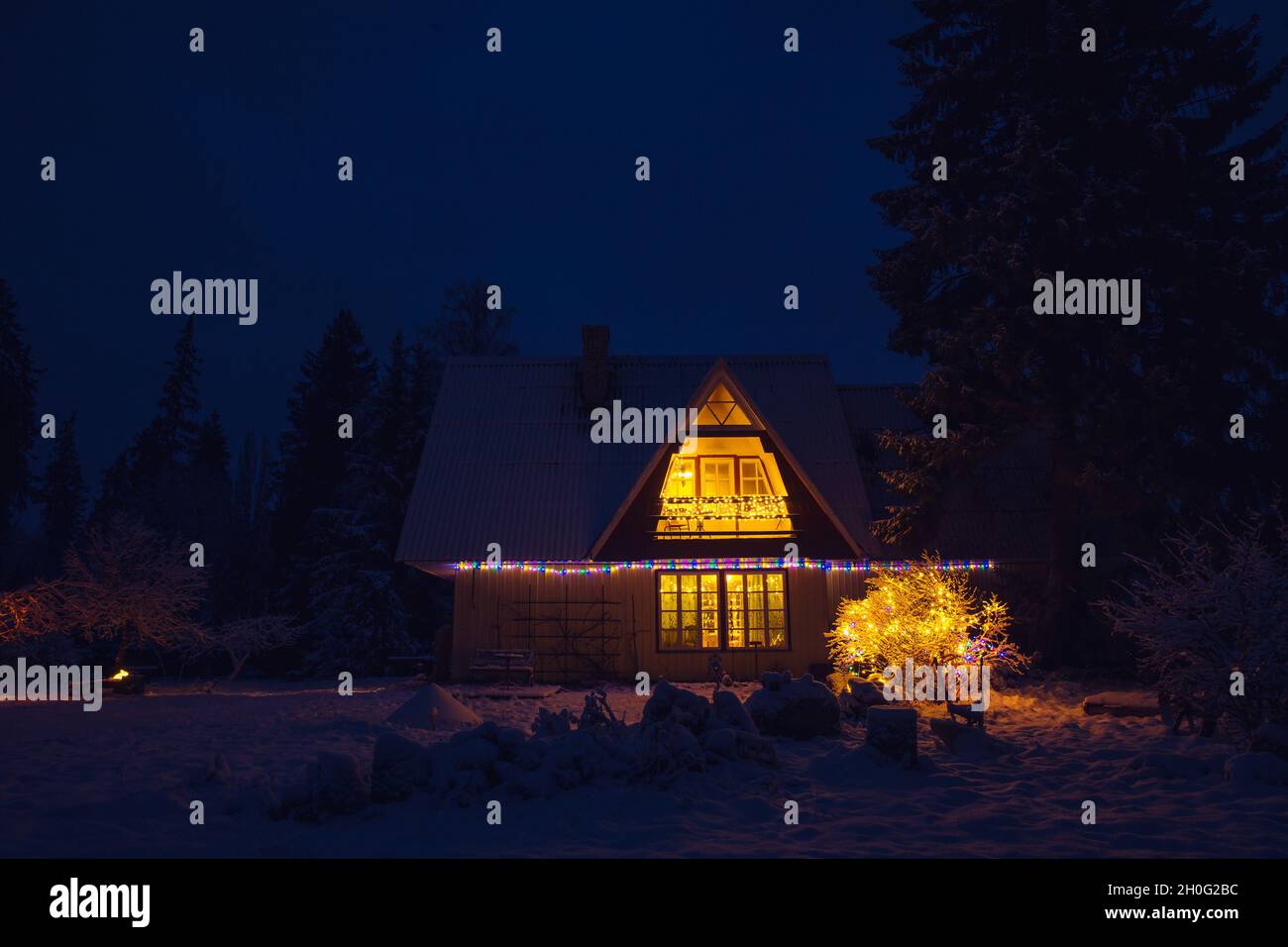 Cute decorated vintage country house home in the middle of forest at night. Tranquil Christmas Eve with snow in garden and light coming from window. Stock Photo