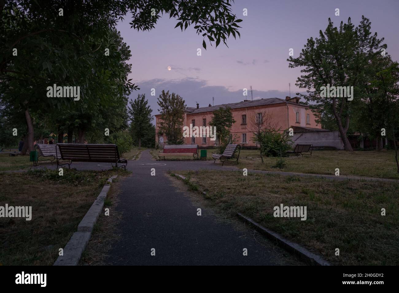 Small town old park plunging in dusk. Outdoor city place for walking with decayed old benches and moon above clouds in dark sky. Stock Photo