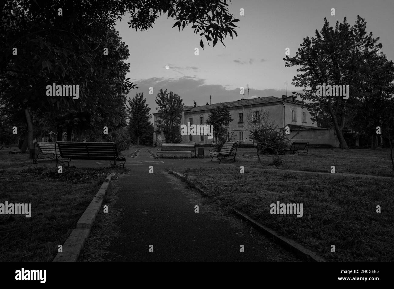 Black and white photo of small town old park plunging in dusk. Outdoor city place for walking with decayed old benches and moon above clouds in dark Stock Photo