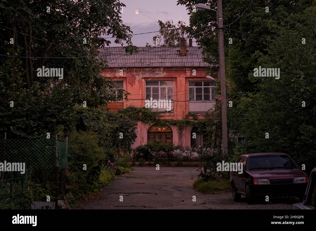 Night outdoor photo of small town old two storey house yard drowned in greenery with an old car parked on the road side in dusk with moon above clouds Stock Photo