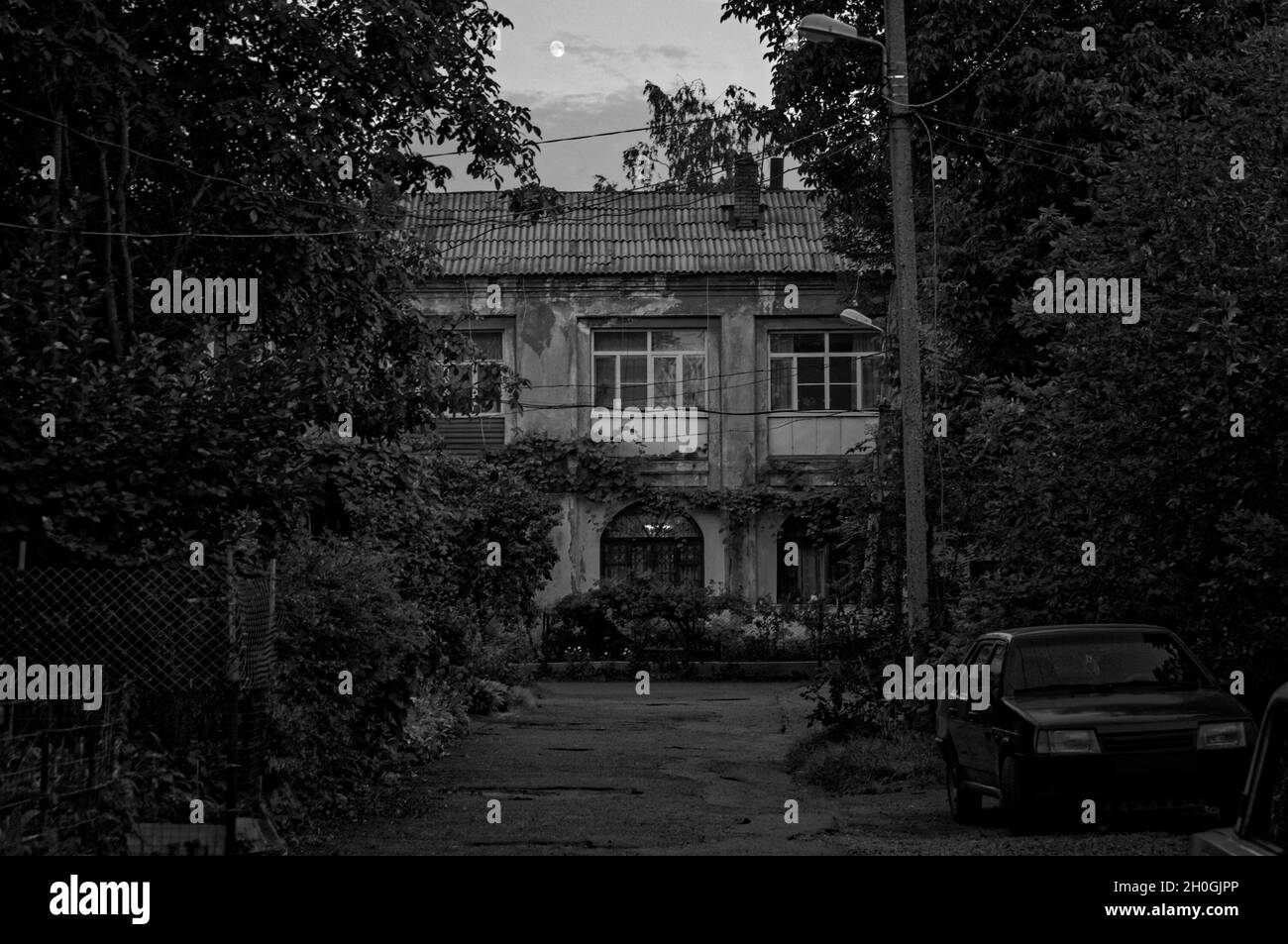 Black and white outdoor photo of a small town old two-story house yard drowned in greenery with an old car parked on the road side in dusk with moon Stock Photo
