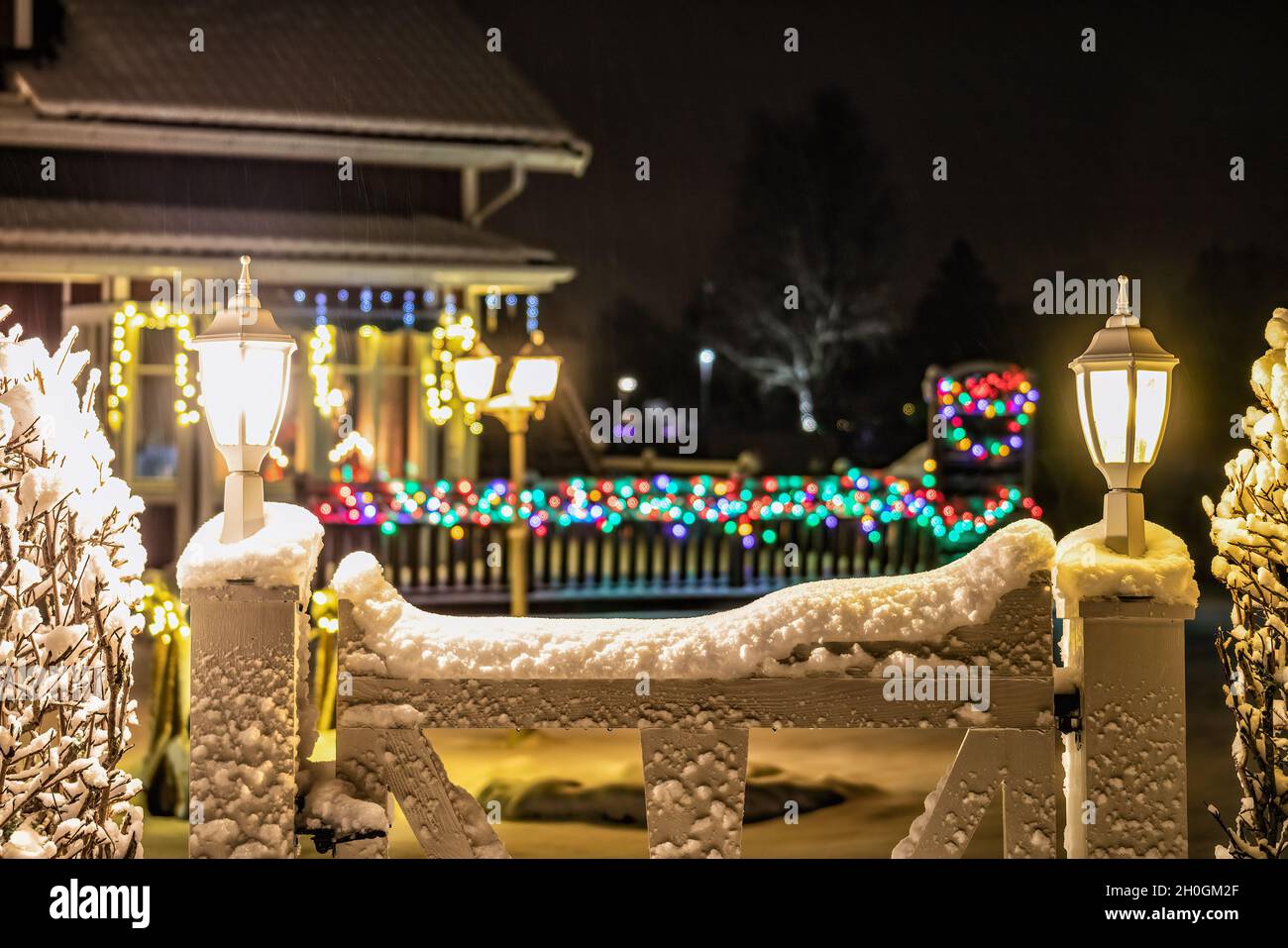 Blurry Christmas light decoration on villa and wooden fence, multicolored lights glowing. White garden gate with two white lanterns at foreground Stock Photo
