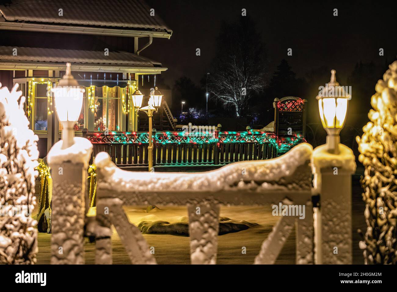 Christmas light decoration on villa and wooden fence, multicolored lights glowing. Blurry garden gate with two white lanterns at foreground Stock Photo