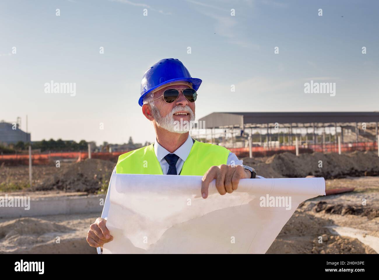 Senior engineer with blueprints standing at building site and looking up in the air Stock Photo