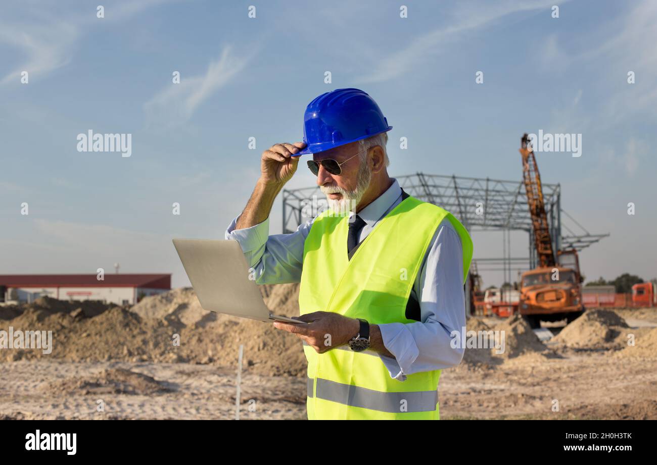 Senior engineer with laptop standing in front of metal construction at building site Stock Photo